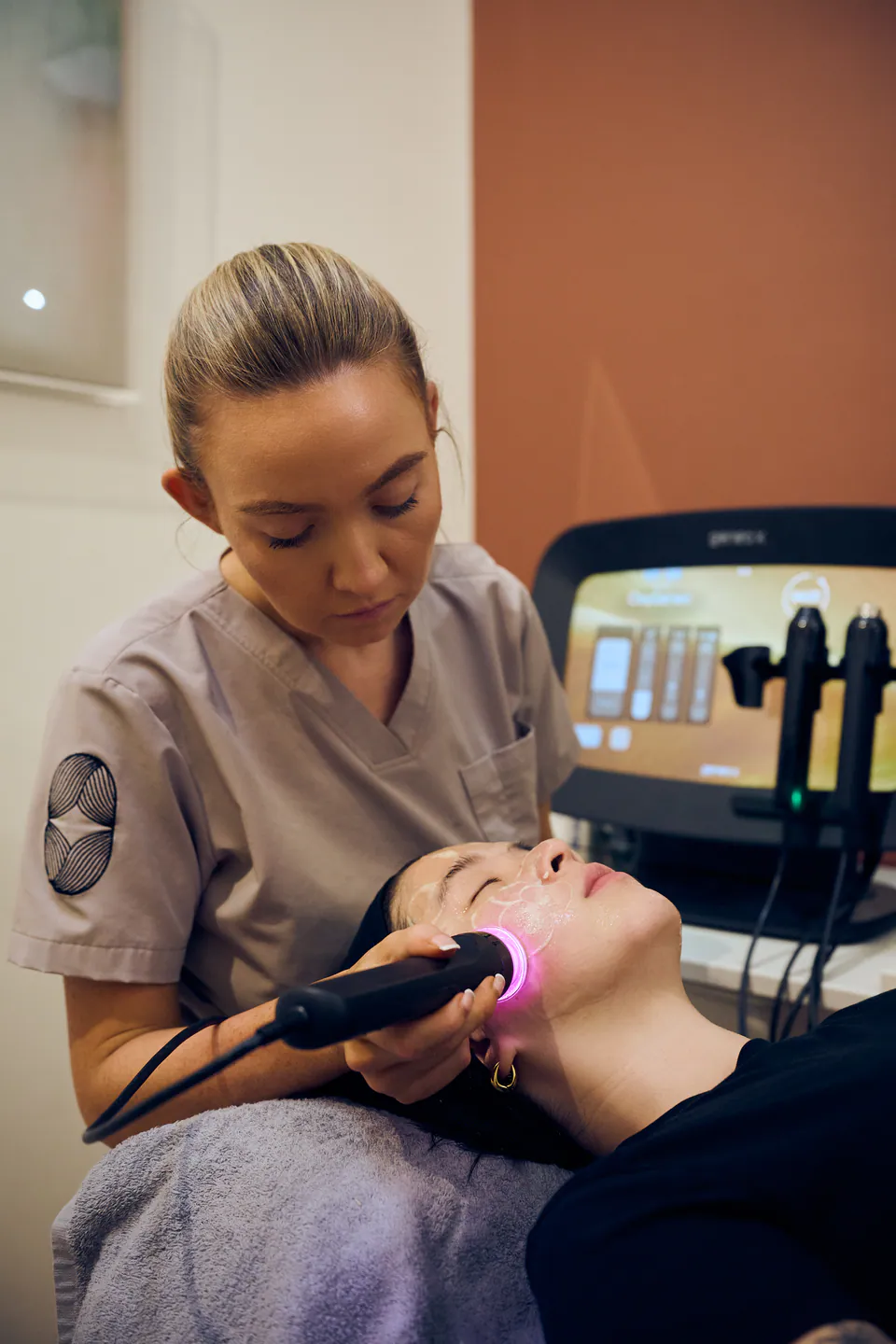 A therapist guiding the Geneo X handpiece along a client's cheek, its light glowing against the skin